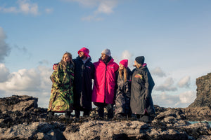A group of people stood on a rock arm in arm looking out to sea and wearing dryrobes