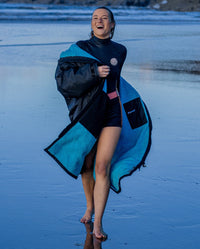 1|Woman running across a beach, wearing a wetsuit and Black Blue dryrobe® Advance Long Sleeve