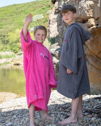 Two boys stood next to a lake, one of them waving, both wearing Kids Towel Ponchos