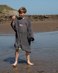 Boy stood on the beach pointing at the sky, wearing Kids Towel Poncho in Slate Grey