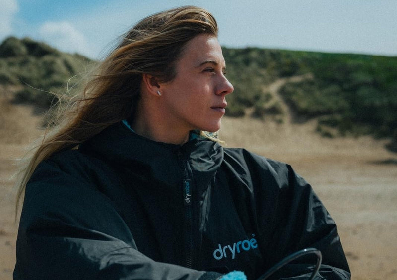 A female surfer sat on beach with a pink and white surfboard on her lap and wearing a black dryrobe 