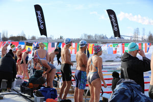 Competitors in colorful swimwear and caps preparing for their heat on the ice-covered pool deck, lined with international flags and Dryrobe® banners.