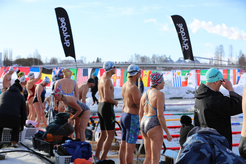 Competitors in colorful swimwear and caps preparing for their heat on the ice-covered pool deck, lined with international flags and Dryrobe® banners.