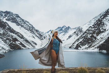 A woman wearing a white dryrobe and swimming costume with snowy mountains and water in the background