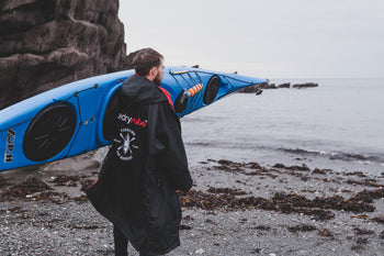 Jack Hampton stood on a beach holding a blue kayak and wearing a dryrobe change robe