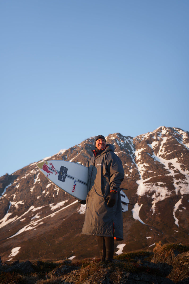 Andrew Cotton wearing a wetsuit and dryrobe holding a surfboard in Norway with snowy mountains behind him