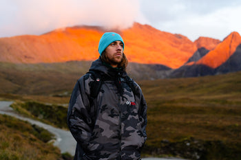 A man stood with a Black Camo Black dryrobe and blue beanie with the Scottish mountains glowing in the sunset behind him 