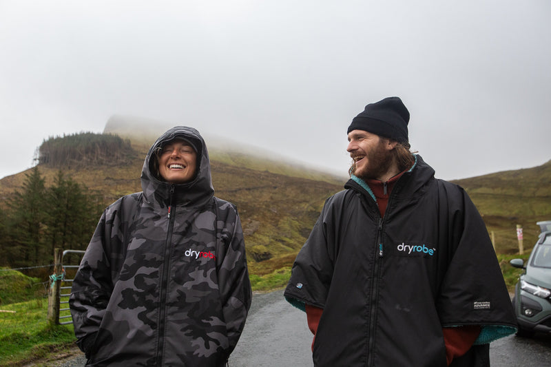 A female and male surfer smiling in dryrobes with the misty Irish countryside in the background 