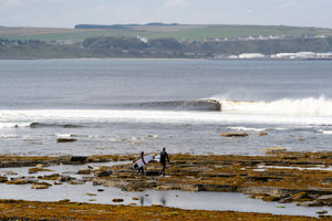 Two surfers walking over rocks to the sea in Scotland wearing hooded wetsuits