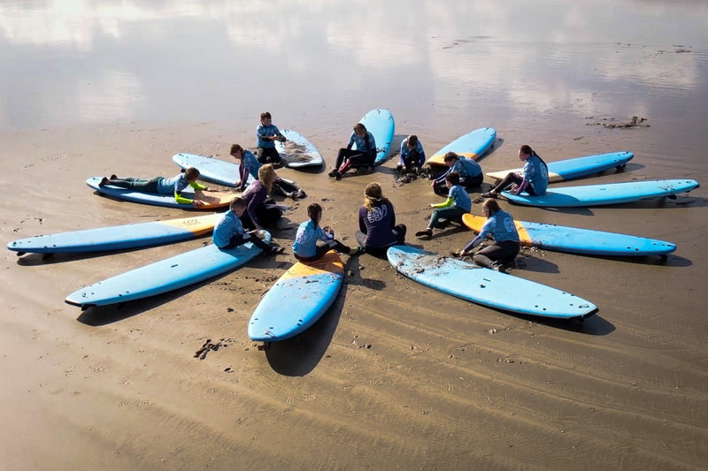 A group of young surfers and instructors sitting on their surfboards in a circle on the sand, engaging in a group discussion during a Liquid Therapy session.