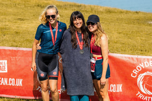 Three women stood together with medals around their necks in front of a red Croyde Ocean Triathlon sign on Baggy Point
