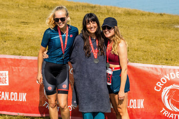 Three women stood together with medals around their necks in front of a red Croyde Ocean Triathlon sign on Baggy Point