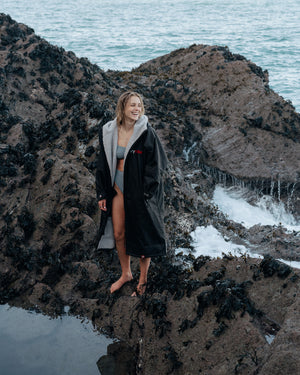 A smiling Sophie Hellyer stood on the edge of a rock pool wearing a dryrobe, with the sea behind her