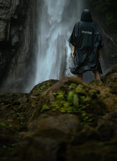 Person wearing a black Dryrobe Lite standing in front of a waterfall.