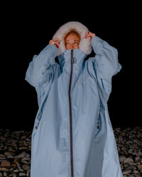 Woman stood on pebble beach at night, wearing dryrobe Advance Long Sleeve Alpine Range change robe in Alpine Ice Blue, pulling up hood