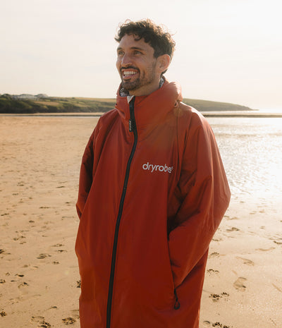 *MALE* A man smiling on a sandy beach wearing a red-orange Dryrobe waterproof jacket. The background shows water and distant land under a bright sky.