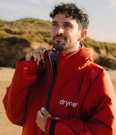 *MALE* A man wearing a bright red Dryre waterproof jacket stands on a sandy beach with dunes in the background. The white 'dryrobe' logo is visible on the left chest of the jacket.