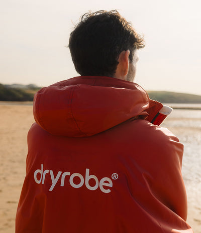 *MALE* A man viewed from behind wearing a red hooded Dryrobe jacket on a sandy beach with water in the background.