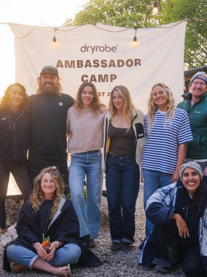 A group of seven people posing outdoors in front of a white banner that reads 'dryrobe AMBASSADOR CAMP'. The group is wearing various casual and outdoor apparel items, with trees and string lights visible in the background.
