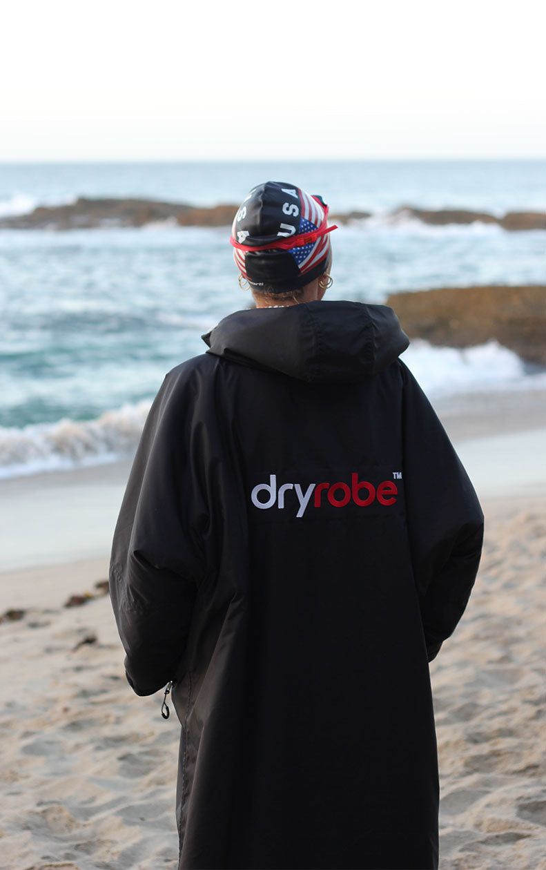 A person standing on a sandy beach facing the ocean, wearing a black hooded Dryrobe garment with the brand name printed on the back in white and red.