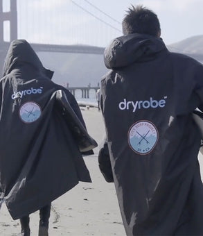 Two people seen from behind wearing dark gray hooded dryrobes with white 'dryrobe' logos and circular patches, walking outdoors on a beach with a bridge and hills in the background.