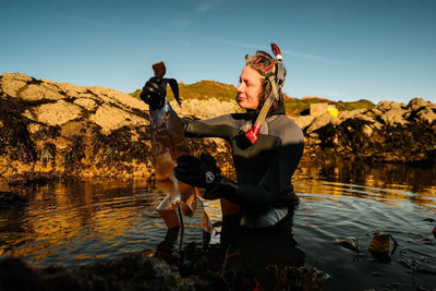 Person in scuba gear holding a small figurine by a body of water with a scenic background.