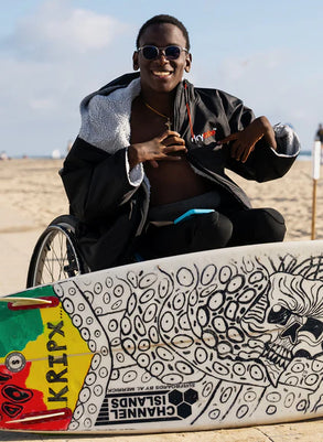 Person in a wheelchair wearing a Dryrobe® change robe with a colorful skateboard on a beach