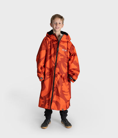 *MALE* A young boy modeling a bright orange and red camouflage hooded raincoat with black zippers and black cuffs, standing against a light gray studio background.