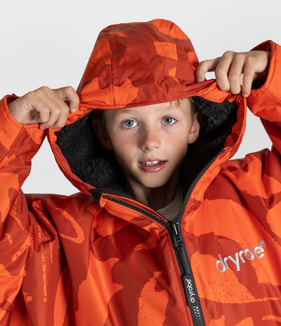 *MALE* A child wearing an orange and red camouflage patterned hooded jacket with a black fleece-lined hood, holding the hood up against a light gray studio background. The brand name 'dryroe' is visible on the chest and zipper pull.