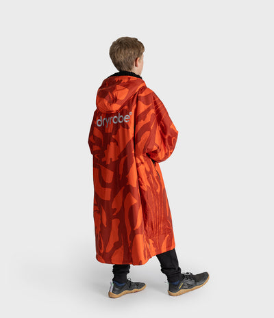 *MALE* A young boy modeling a bright orange and red camouflage hooded raincoat with black zippers and black cuffs, standing against a light gray studio background.