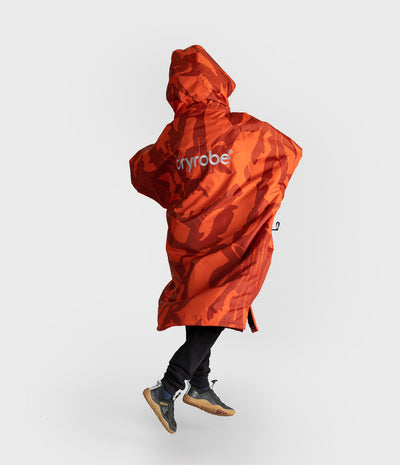 *MALE* A person wearing a bright orange and red camouflage hooded rain poncho with the brand name 'dryrobe' printed in white on the back, paired with black pants and gray sneakers, captured mid-step against a light gray studio background.