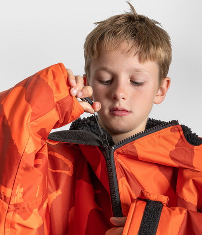 *MALE* A close-up of a young boy wearing a bright orange and red camouflage winter jacket with black fleece lining and a black zipper, shown against a light gray studio background.