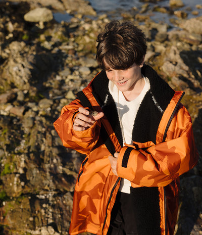 *MALE* A boy wearing a bright orange camouflage-patterned waterproof jacket with black fleece lining, standing outdoors on a rocky shoreline.