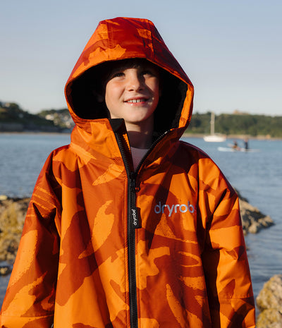 *MALE* A smiling child wearing a bright orange and brown camouflage hooded rain jacket with a black zipper and the brand name 'dryrob' on the chest, standing outdoors near water and rocks.