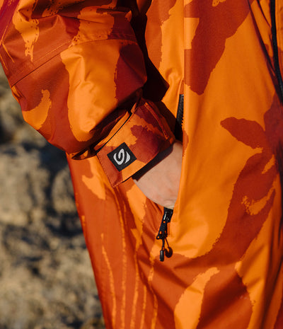 *MALE* Close-up of a person wearing a bright orange and dark red camouflage-patterned jacket, with a hand in a zippered pocket and a small black logo patch on the cuff.