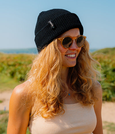 A smiling woman outdoors wearing a black knit beanie with a small white logo, round amber-tinted sunglasses, and a light beige ribbed tank top. The background shows blurred greenery and blue sky.