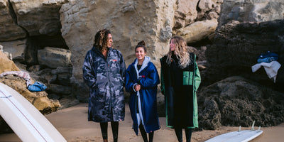 Three people standing on a beach with surfboards, wearing colorful dryrobes.