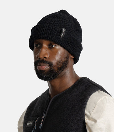 A man wearing a black knit beanie with a small white 'dryrobe' brand tag and a black fleece vest over a cream shirt, photographed against a light gray studio background.