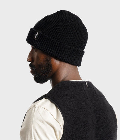 A man in profile wearing a black ribbed knit beanie with a small white logo patch on the cuff, paired with a cream shirt and a black fleece vest, against a light gray studio background.
