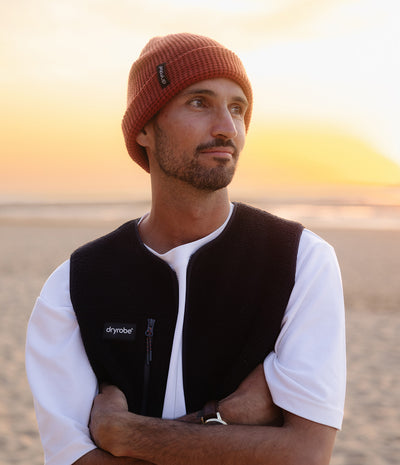A man wearing a rust-colored knit beanie and a black Drylab fleece vest over a white t-shirt, standing on a sandy beach at sunset.