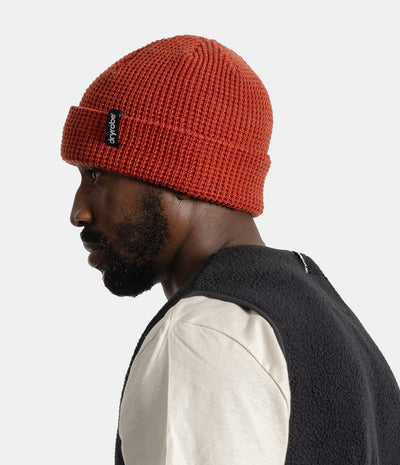 A man in profile wearing a textured orange-red knit beanie with a black Fly London logo patch on the cuff, paired with a cream shirt and a dark fleece vest, against a light gray studio background.