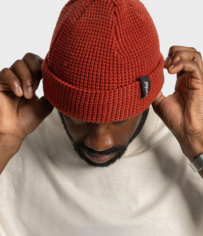 A man wearing a rust-orange ribbed knit beanie hat with a small black brand tag, adjusting the brim with both hands against a light gray background.