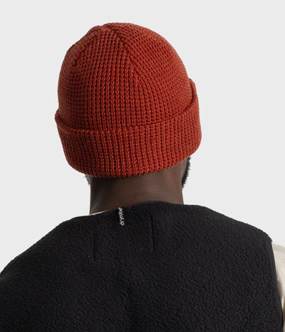 A person shown from the back wearing a rust-orange ribbed knit beanie and a black fleece vest against a light gray studio background.
