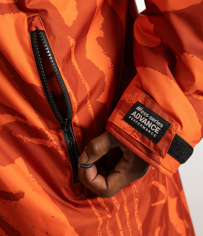 *MALE* Close-up of an orange patterned Dryrobe with a black zipper and a visible sleeve patch reading 'no-series ADVANCE PERFORMANCE'. A hand is shown zipping the jacket.