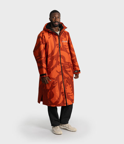 *MALE* A man modeling a long orange hooded Dryrobe with an abstract dark red-orange pattern, featuring a white 'swoosh' logo on the chest, black zippers, and black cuffs, paired with black pants and beige sneakers against a light gray background.