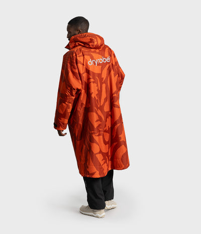 *MALE* A model shown from the back wearing a long orange hooded Dryrobe with a tonal abstract pattern and the brand name 'dryrobe' printed in white on the upper back, paired with black pants and light-colored sneakers against a light gray studio background.