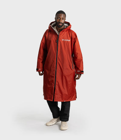 *MALE* A man wearing a long red hooded Dryrobe jacket with black zippers, black pants, and beige sneakers, standing against a light gray studio background.