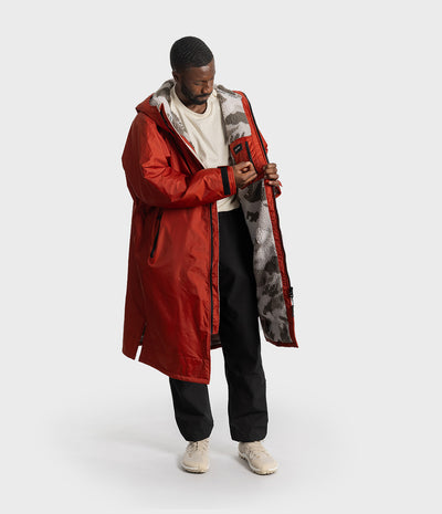 *MALE* A man wearing a long red hooded parka with a gray and black camouflage-patterned interior lining, styled with a white t-shirt, black pants, and beige sneakers, set against a plain light gray studio background.