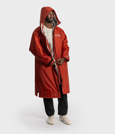 *MALE* A man wearing a hooded red jacket with a white graphic print on the chest, layered over a white t-shirt, black pants, and beige sneakers, standing against a light gray studio background.
