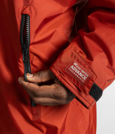*MALE* Close-up of a person wearing a bright red technical jacket, pulling a black zipper on the chest while showing a branded cuff label that reads 'MAXI-SERIES ADVANCE PERFORMANCE'.